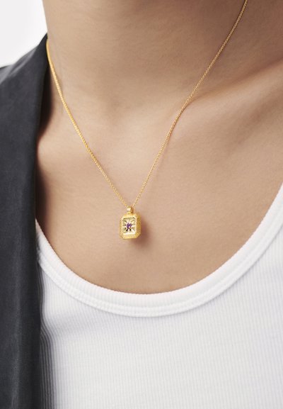 Kuldne kaelakee ristküliku kujulise riputiga, millel on lilla vääriskivi, tekstureeritud disain ja peen kett, toetub valgele soonilise pinna valmistatud topile.