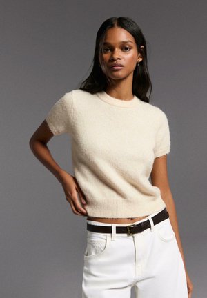 Nuori nainen, jolla on pitkät tummat hiukset, pukeutuneena lyhythihaiseen kermaan sävytettyyn neulepaitaan, valkoisiin housuihin ja mustaan vyöhön, poseeraa harmaata taustaa vasten.