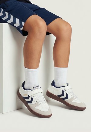 Barn iført hvide sneakers med marineblå chevronstriber, hvide sokker og marineblå shorts, der sidder på en hvid blok mod en ensfarvet baggrund.