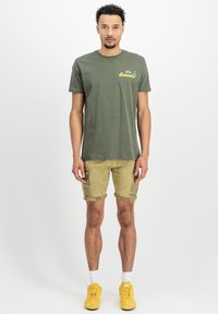 Oliven grøn t-shirt i bomuld med "Hawaii" grafik, parret med khaki cargo shorts med lommer; modellen har gule sneakers på.