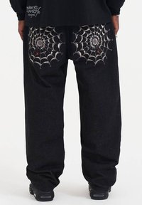 Zwarte denim broek met geborduurde spinnenwebontwerpen op de achterzakken, met zilveren draden en kleine rode accenten. Losse pasvorm.