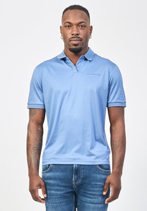 Polo shirt azzurro chiaro con colletto, maniche corte e logo sul petto a sinistra. Il tessuto appare morbido con una vestibilità rilassata, abbinato a jeans blu.