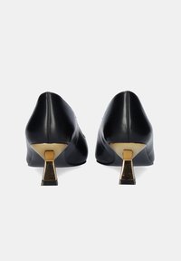 Zapatos de cuero negro con punta afilada, que cuentan con un acento metálico dorado en un tacón esculpido. Textura suave, diseño elegante y sin patrones visibles.