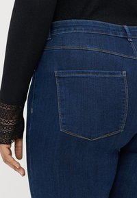 Jeans de ganga azul escuro com um bolso traseiro, apresentando uma cintura média e uma textura suave. Acompanhados por uma blusa preta de manga longa com renda.