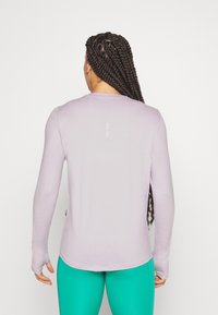 Langærmet lys lilla bluse med rund kant og hul til tommelfingeren, parret med grønne leggings. Subtil logo-detalje på ryggen. Glat tekstur.