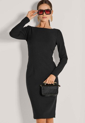 Woman in a fitted black long-sleeve dress holding a small black handbag and adjusting large red-tinted square sunglasses.