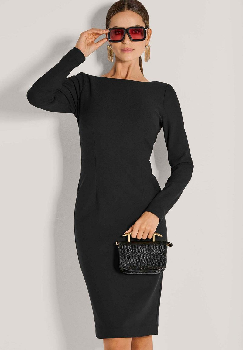 Frau in einem enganliegenden schwarzen Langarmkleid, die eine kleine schwarze Handtasche hält und große rotgetönte quadratische Sonnenbrillen anpasst.