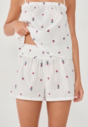 Femme portant un débardeur en coton blanc et un short avec de petits motifs floraux roses et verts, levant légèrement le débardeur.