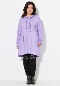 Lila hooded tuniek met voorzakken en tekstversieringen, gecombineerd met zwarte broek met witte tekst en gestudeerde zwarte laarzen.