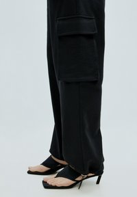 Zwarte cargobroek met een zijzak, gemaakt van zacht materiaal, gecombineerd met elegante zwarte open-toe haksandalen.