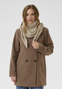 Brun uldfrakke med dobbeltknapper og sidelommer, parret med et lys beige tørklæde, præsenteret mod en ensfarvet baggrund.
