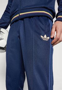 Marineblauwe Adidas trainingsbroek met een gestructureerde afwerking, voorzien van een gouden logo, accentstrepen en zijzakken. Een voetbal wordt in één hand vastgehouden.