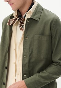 Olivengrüne Canvas-Jacke mit einem Kragen, Vordertasche und schwarzen Knöpfen, getragen über einem blassbeigen Hemd. Brauner Schal mit Muster um den Hals.