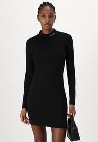 Melna garām piedurknēm kleita ar augstu apkakli un Calvin Klein logo. Pieguļošs dizains, gluda tekstūra un ceļa garumā. Modelis tur mazu melnu somiņu.