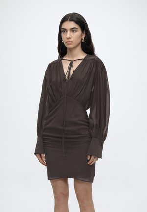 Vrouw draagt een donkerbruine mini-jurk met lange mouwen, doorschijnende pofmouwen en een strik bij de diepe V-hals, staand tegen een effen achtergrond.