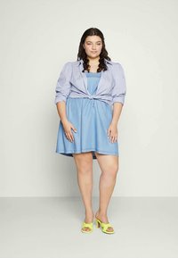Vestido de ganga azul claro com corpete ajustado e saia evasé, combinado com uma camisa azul clara atada. Usado com saltos altos amarelos brilhantes.