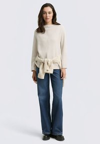 Cremefarbenes, geripptes Oberteil mit langen Ärmeln, Taillendetail mit Bindung, kombiniert mit dunkelblauen, ausgestellten Jeans und schwarzen Stiefeletten. Einfaches, lässiges Outfit.