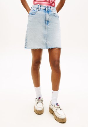 Person iført en lyseblå denimnederdel, hvide sokker, kraftige hvide sko og en rød- og hvidstribet top med hænderne bag ryggen.