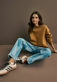 Donna con lunghi capelli castani seduta sul pavimento contro un muro marrone, che indossa un maglione senape, jeans a pezzi blu e sneakers bianche con strisce nere.