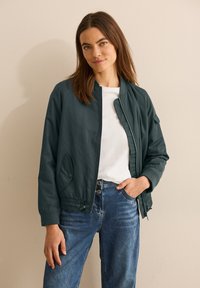 Giubbotto bomber verde scuro con colletto e polsini a costine, dotato di zip e tasche laterali, indossato sopra una camicia bianca e jeans blu.