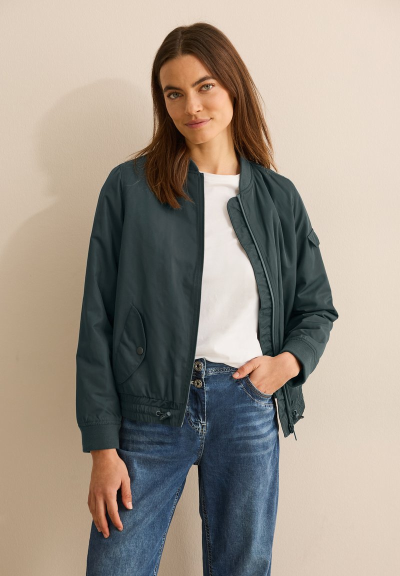 Giubbotto bomber verde scuro con colletto e polsini a costine, dotato di zip e tasche laterali, indossato sopra una camicia bianca e jeans blu.