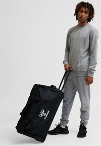 Czarna torba podróżna na kółkach z uchwytami, ozdobiona białym logo, obok osoby w szarej bluzie i spodniach dresowych oraz czarnych trampkach.