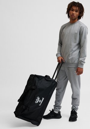 Zwarte draagtas op wieltjes met handvatten en een wit logo, naast een persoon die een grijze trui en joggingbroek en zwarte sneakers draagt.