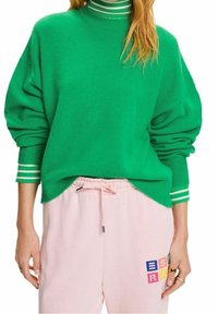 Femme portant un pull vert avec des poignets et un col rayés, associé à un pantalon rose clair à cordon avec un motif coloré en lettres type bloc.