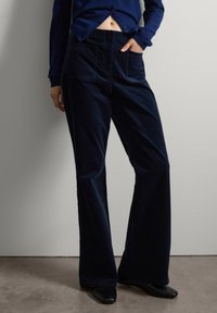 Persona vistiendo pantalones anchos de pana azul marino, con una mano en el bolsillo, combinados con un cárdigan azul marino abotonado y zapatos de punta negros.