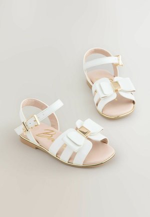 Valged lakitud sandaalid, millel on lipsukujuline detail, kuldsed aktsendid ja pahkluu rihm. Tasane talla osa ja sile beež sääreosa.