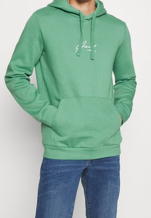 Mann trägt grünen Kapuzenpullover mit Fronttasche und weißem kursive Schriftzug, kombiniert mit blauen Jeans, Hände in den Taschen.