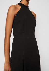 Vestido negro de cuello halter hecho de tela suave; torso ajustado con una cintura definida; falda acampanada con pliegues.
