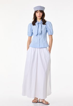 Vrouw draagt een lichtblauwe gestreepte blouse met pofmouwen en strik, witte lange rok met zakken, gehaakte hoed en witte sandalen, rechtopstaand.