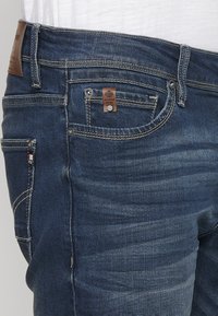 Tamno plavi traperice s smešnim kožnim dodatkom, dizajn sa pet džepova i vidljivim detaljima šivanja. Ima prednji džep s akcentom od hardvera.