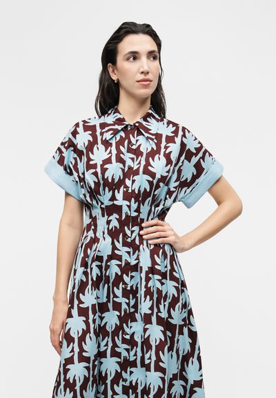 Γυναίκα που φοράει καφέ φόρεμα με μεγάλα ανοιχτό μπλε μοτίβα φοίνικα, με κοντά, γυρισμένα μανίκια και μυτερό γιακά, στέκεται με το ένα χέρι στη μέση.