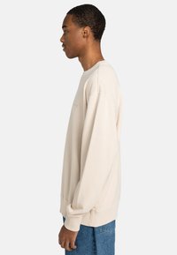 Sudadera beige con cuello redondo, hombros caídos y puños de canalé. Combinada con jeans de mezclilla azules, mostrada en vista lateral.
