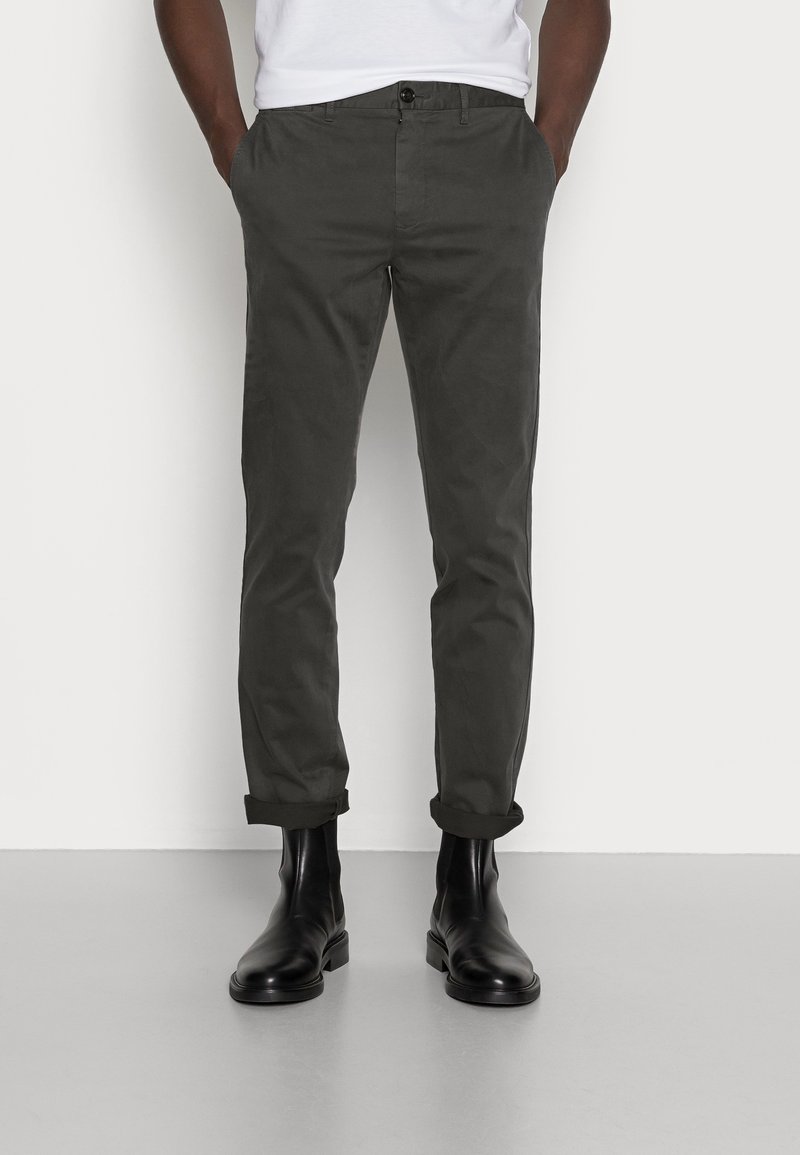 Mężczyzna w podwiniętych ciemnoszarych spodniach i czarnych skórzanych botkach za kostkę, stojący na tle jednolitej białej ściany i szarej podłogi.