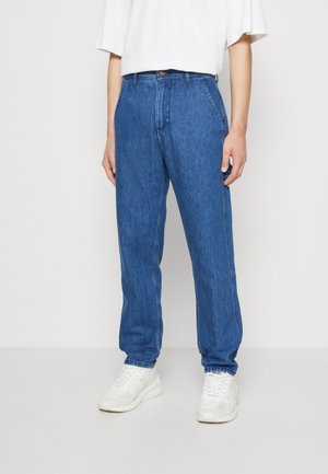Person trägt hoch taillierte, lockere blaue Jeans mit Fronttaschen und weißen Sneakers und steht vor einem einfarbigen Hintergrund.