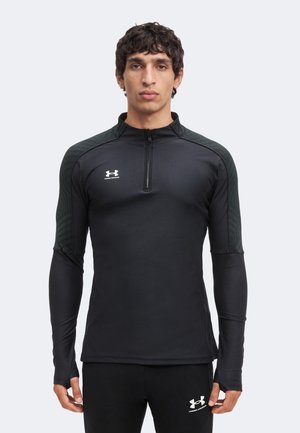 Homme portant un haut de sport noir à manches longues avec fermeture éclair au quart et un pantalon noir assorti, tous deux arborant des logos blancs Under Armour.