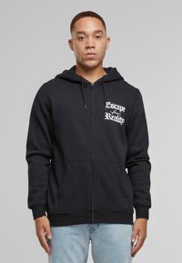 Sort zip-up hættetrøje med en kengurulomme, snor i hætten og hvid tekst "Escape from Reality" på brystet. Blød stofoverflade.