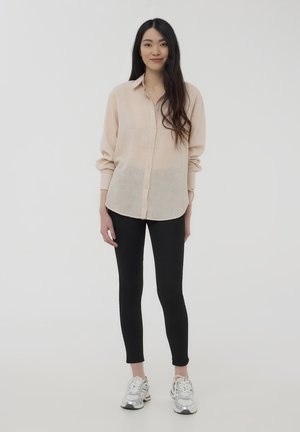 Camicia button-up beige chiaro con maniche lunghe, abbinata a leggings neri e sneaker argentati. Tessuto liscio e vestibilità rilassata.