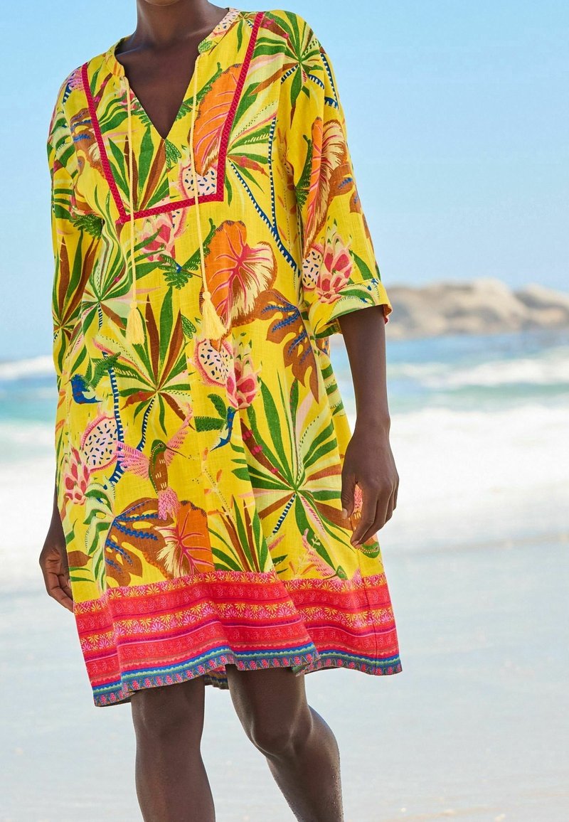 Personne portant une robe jaune ample avec des motifs colorés de feuilles tropicales et floraux, debout sur une plage avec des vagues de l'océan en arrière-plan.