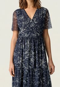 Žena v tmavě modrých šatech s bílými abstraktními vzory, průsvitnými krátkými rukávy a zavinovacím výstřihem do V.