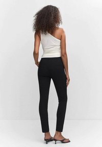 Pantalons noirs ajustés et un top blanc à une épaule, présentant une texture lisse et un design simple. Le modèle est de dos par rapport à la caméra.