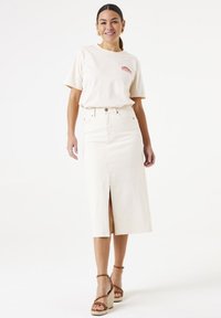 T-shirt en coton beige clair à manches courtes avec un petit logo rouge, associé à une jupe en denim beige taille haute avec fente avant et fermeture à boutons.