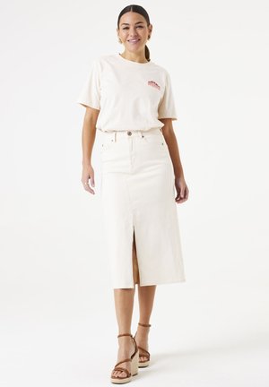 T-shirt en coton beige clair à manches courtes avec un petit logo rouge, associé à une jupe en denim beige taille haute avec fente avant et fermeture à boutons.