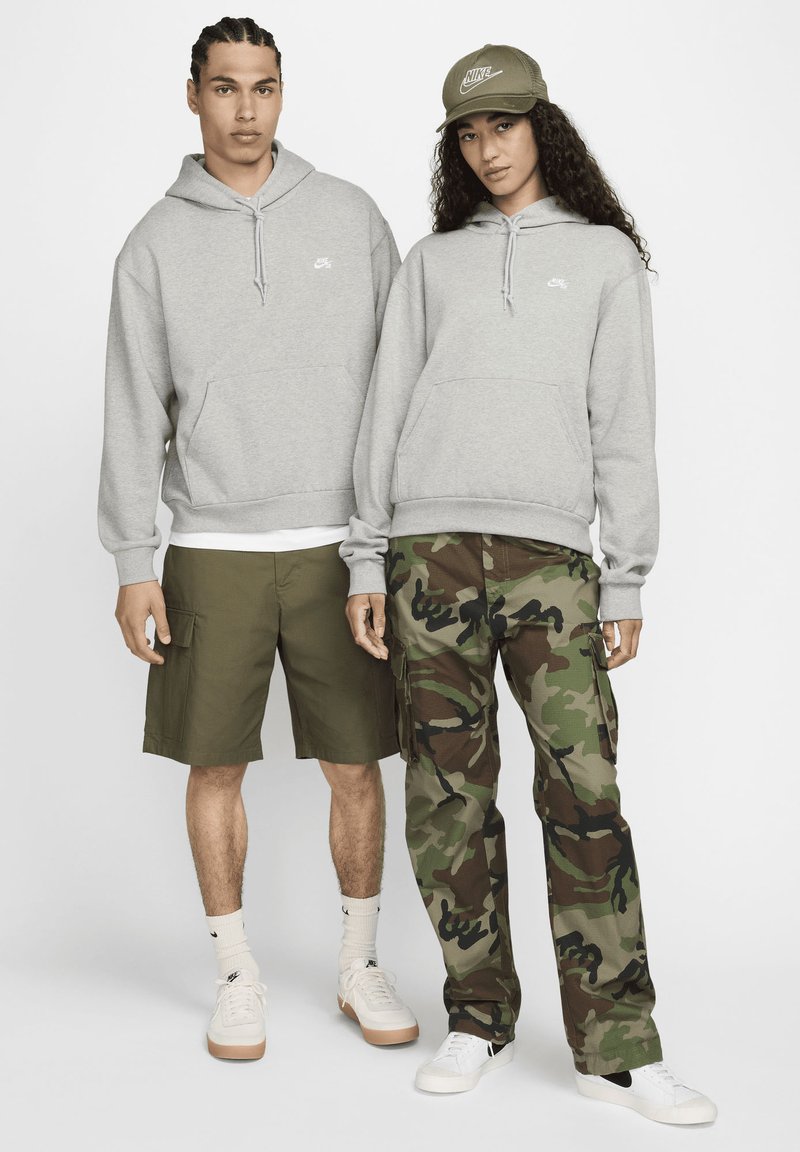 To modeller har på seg en lys grå hettegenser med frontlomme og Nike-logo, kombinert med olivengrønne shorts og kamuflasje cargo-bukser.