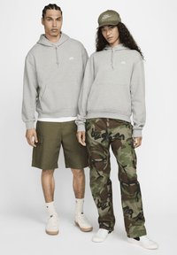 Dva modela nosita ujemajoči se sivi kapucarji z vrvico in žepi spredaj. Eden nosi olivno zelene cargo kratke hlače, drugi maskirne hlače. Oba imata superge in kape.