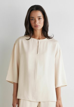 Kvinde med mellemlangt mørkt hår iført løs, beige, kortærmet top med en lille frontslids og matchende bukser foran en ensfarvet baggrund.