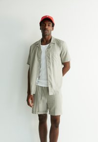 Lichtgrijs shirt met korte mouwen en een zak, gecombineerd met bijpassende korte broek. De persoon draagt een witte tanktop eronder en een rode pet.